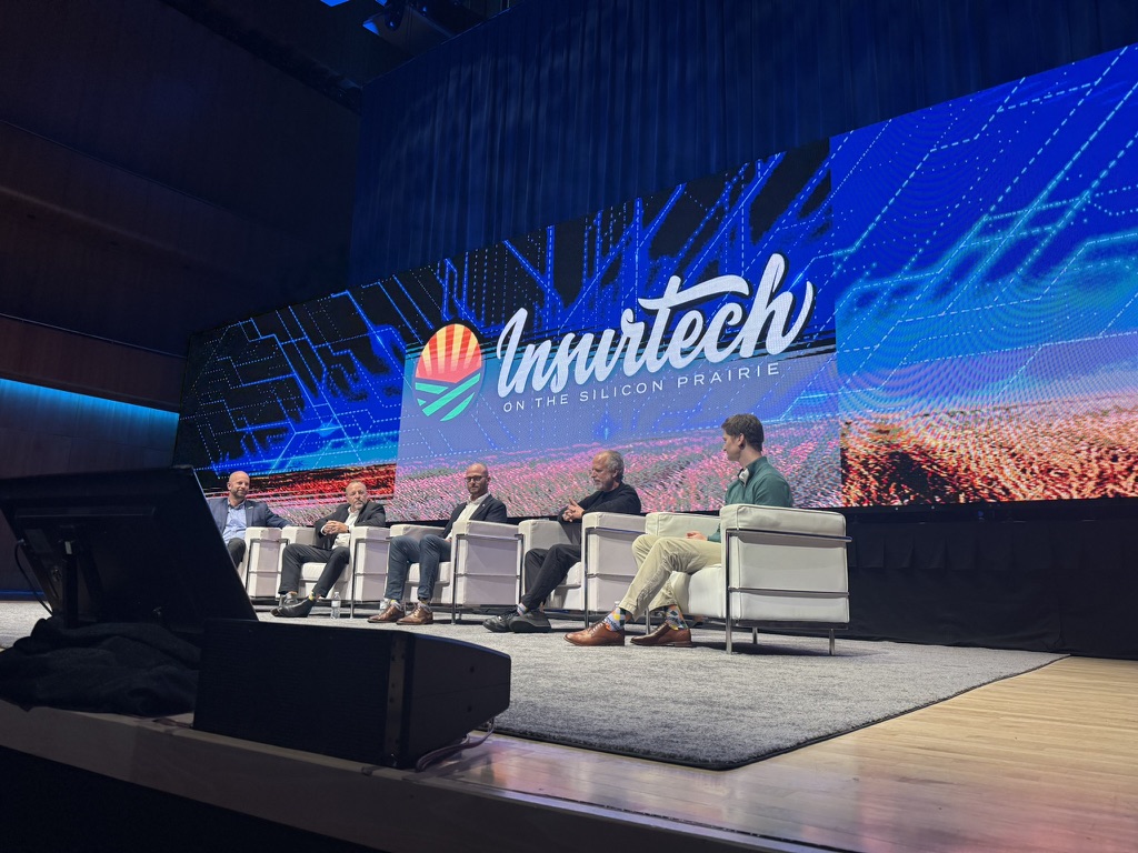 Wide-angle view of five panelists participating in “The Human Element of AI Adoption” at Insurtech on the Silicon Prairie 2026 in Omaha, seated onstage beneath a large illuminated conference screen.
