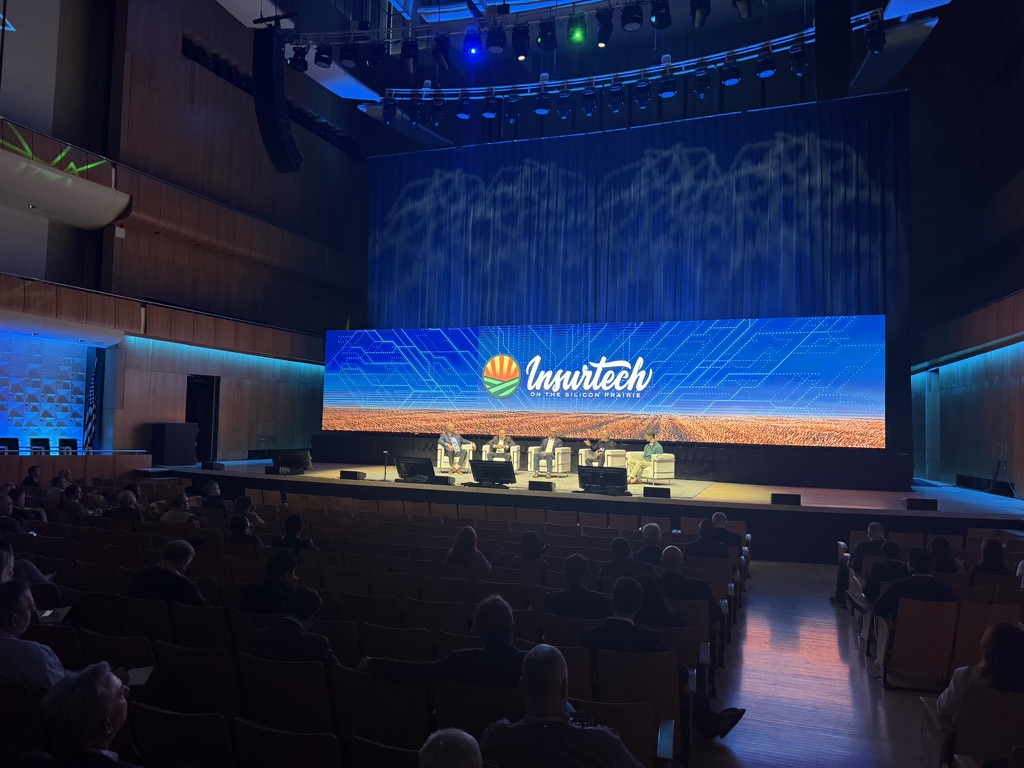Wide view of “The Human Element of AI Adoption” panel at Insurtech on the Silicon Prairie 2026, showing five speakers onstage beneath the conference’s illuminated screen and an audience seated inside the Holland Performing Arts Center in Omaha, Nebraska.