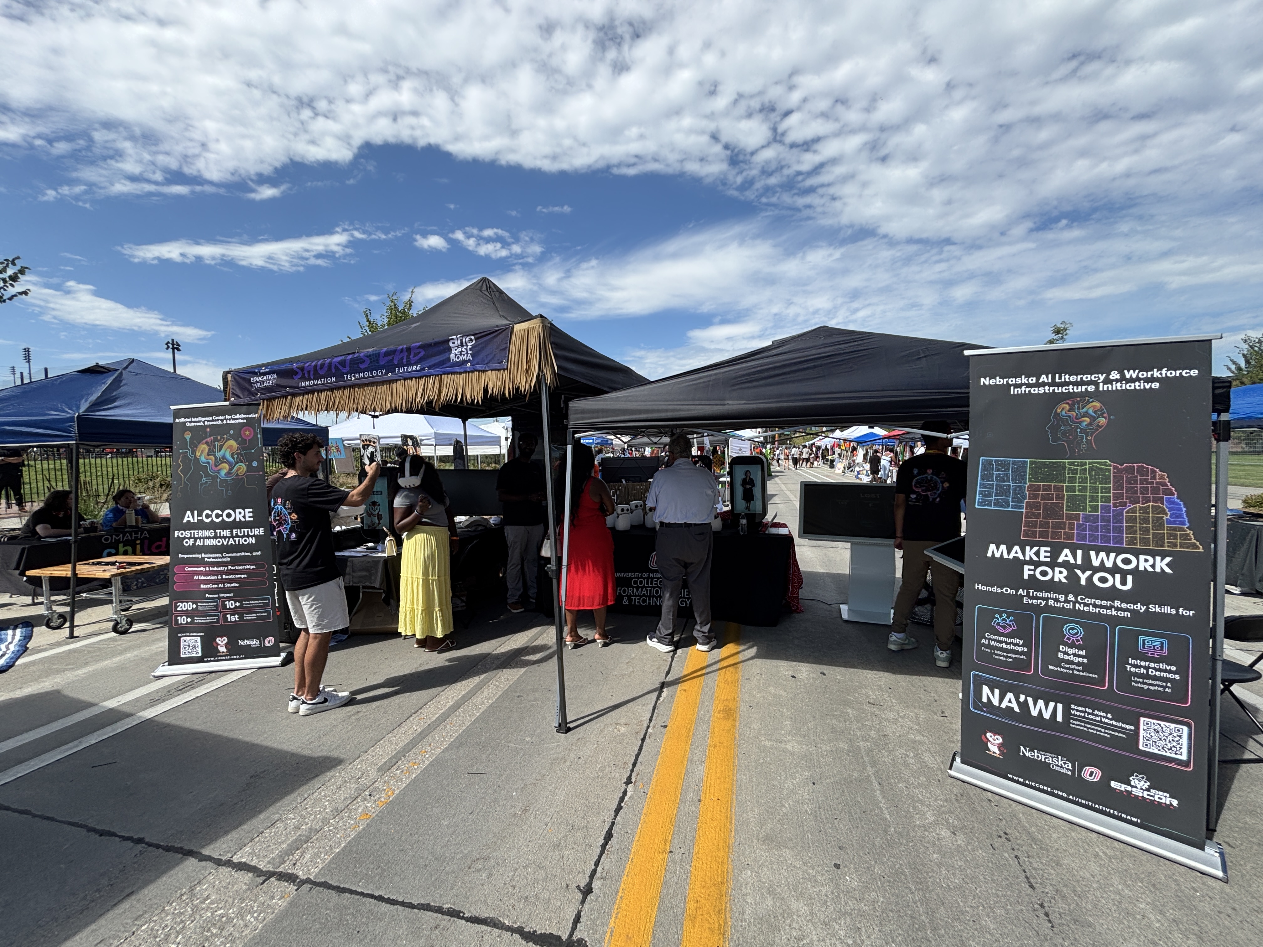 Wide view of the AI-CCORE canopy at Afro Fest Omaha, with tall AI-CCORE and NA'WI retractable banners standing at either end of the tent perimeter and visitors at the tables between them.