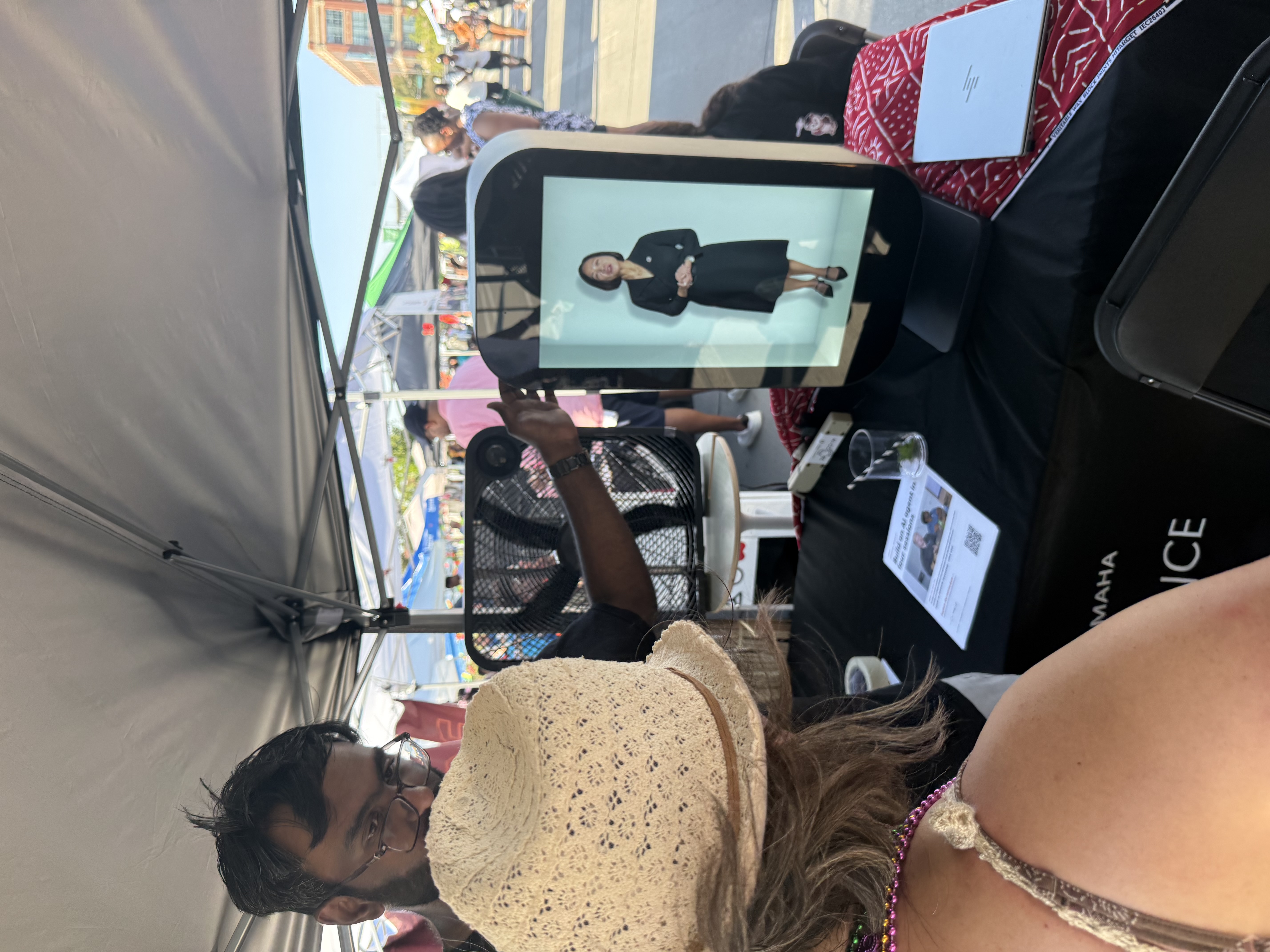 An attendee in a straw hat watches a recorded briefing from Chancellor Joanne Li displayed inside a lit Proto holographic unit under an outdoor festival tent.