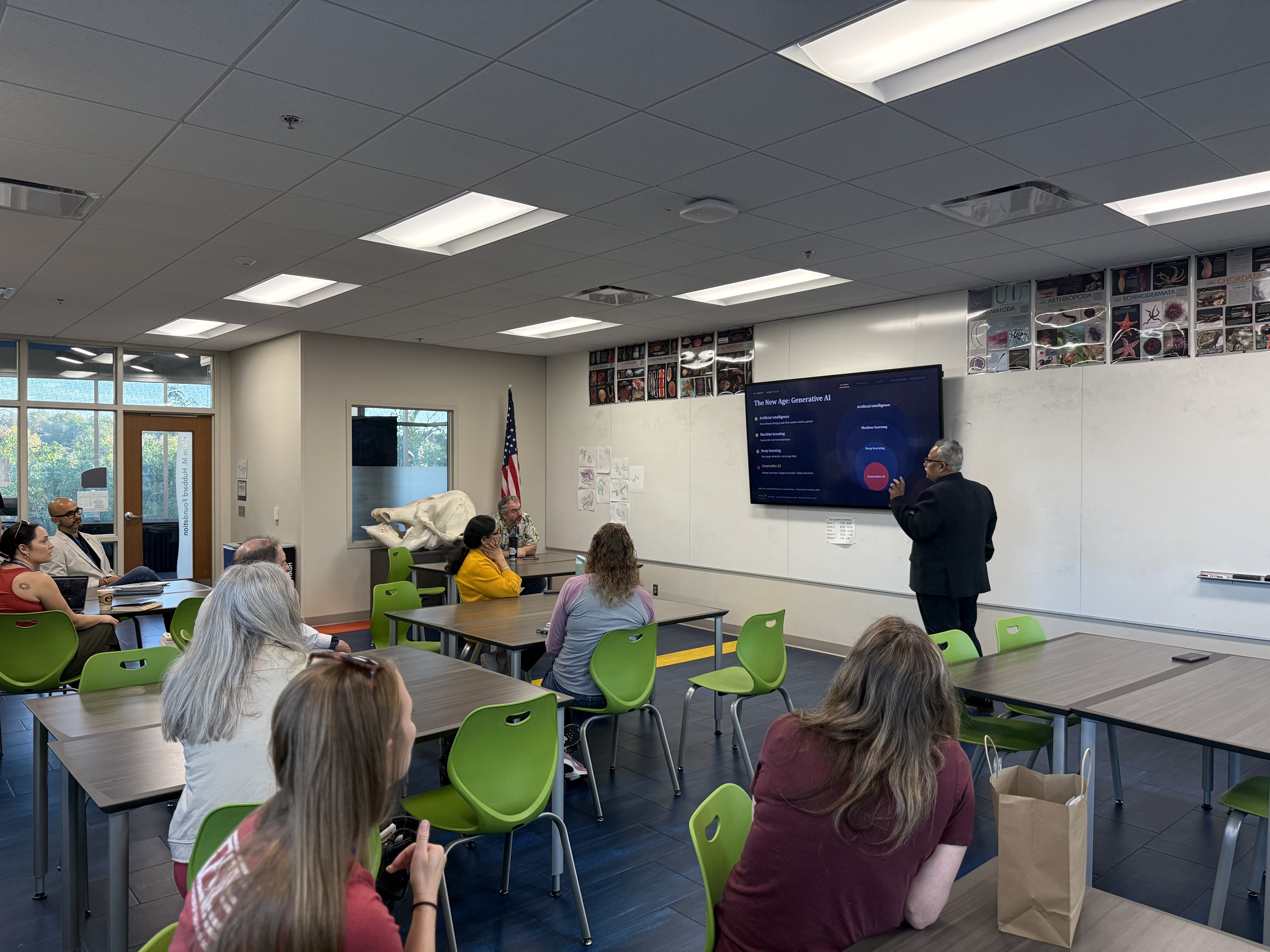 A presenter speaks to a small group of conference attendees seated at tables in a classroom while a wall-mounted screen displays a slide about generative AI.