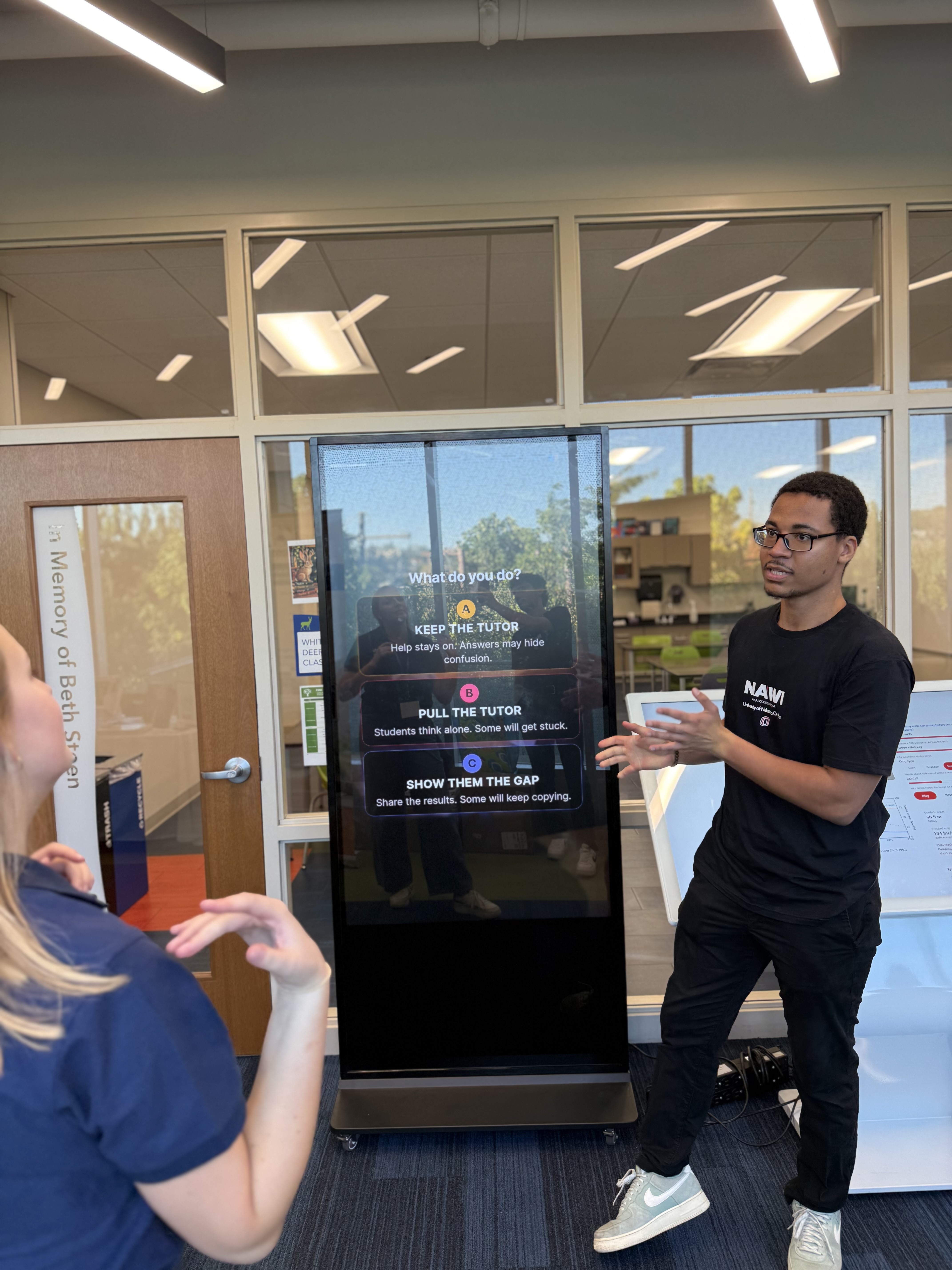 An AI-CCORE team member in a black NA’WI shirt speaks with an attendee beside a tall interactive screen displaying options for an AI tutoring scenario.