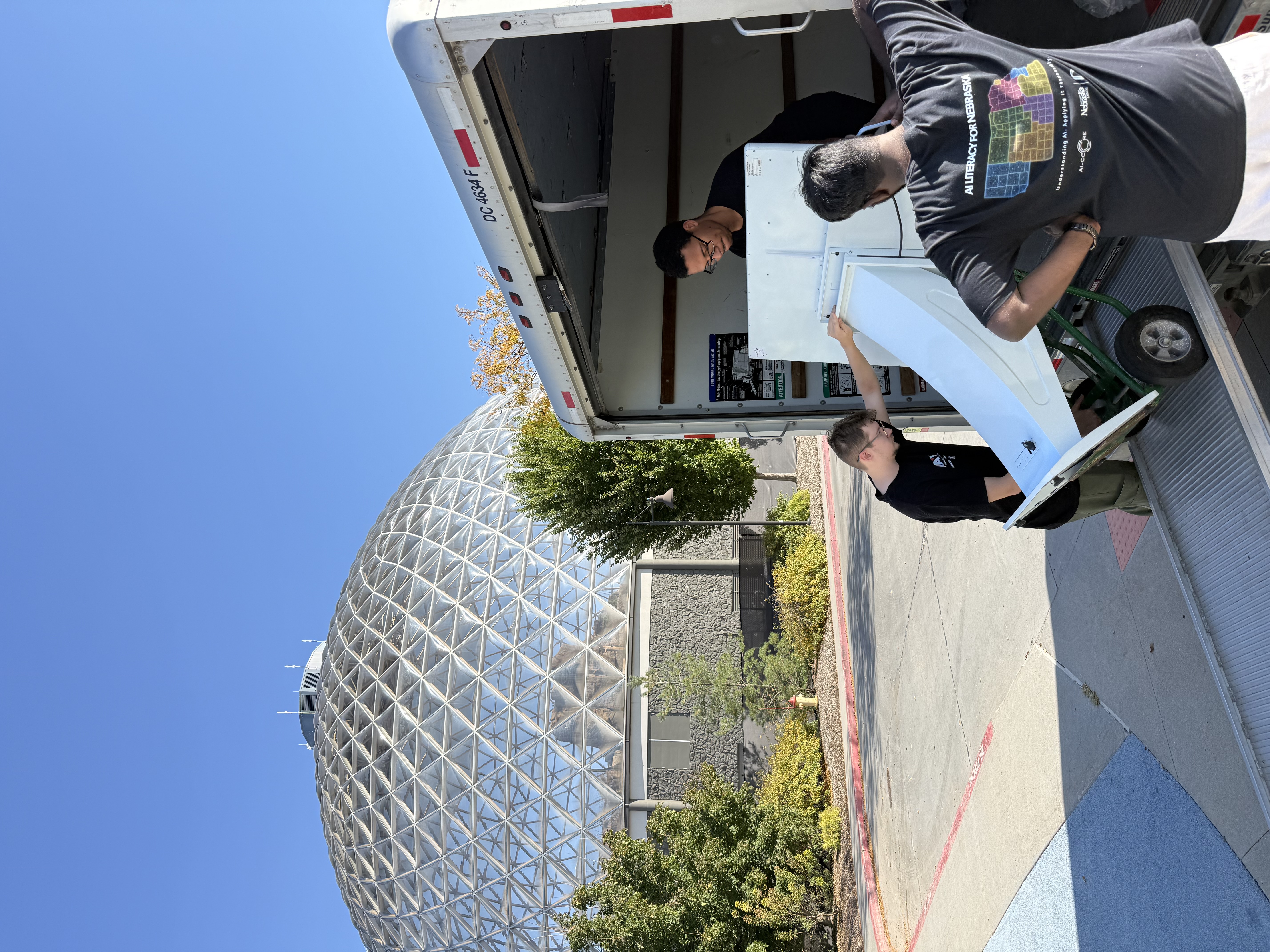 Three team members unload a large white touchscreen kiosk from a moving truck outside Omaha’s Henry Doorly Zoo and Aquarium, with the zoo’s geodesic dome visible in the background.